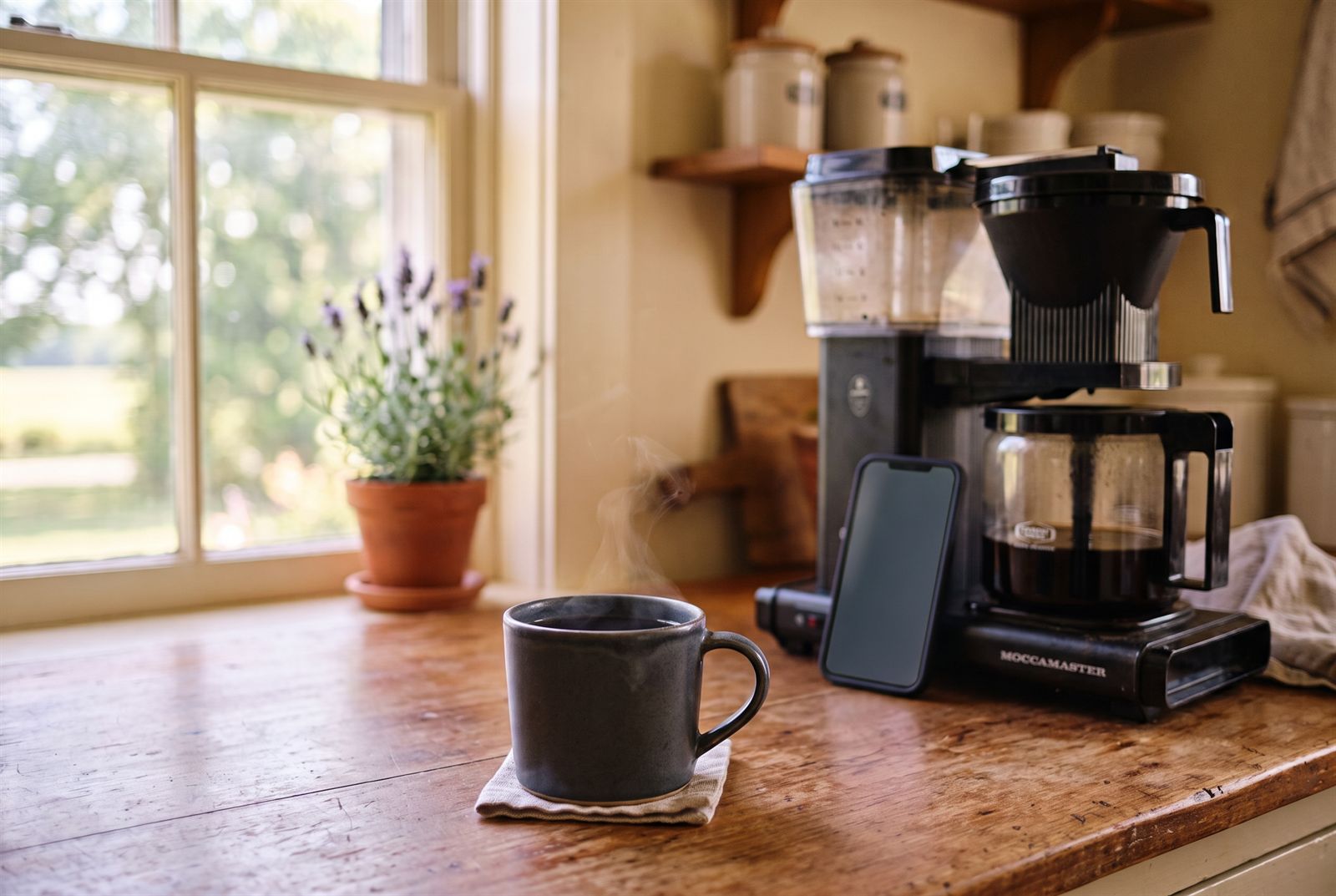 Photo of a phone propped on a kitchen counter in early morning light, next to a full mug of black coffee